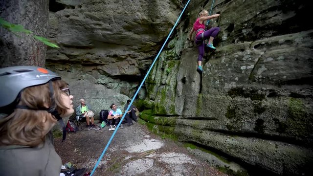 Mature woman climbs up a rock face while searching for places to put her hands and feet as her family looks on.