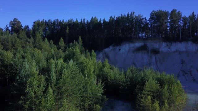 Aerial epic drone flight up from abandoned kaolin quarry with turquoise water, sandy cliffs with fallen trees on the background, sunny evening in anthropogenic landscape of South Ural, Russia