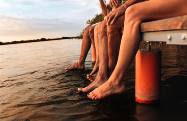 Four young friends sitting on pier with their legs hanging down to the water on a summer day.