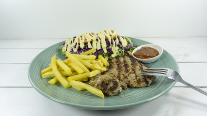 Pork steak in a dish on a white wood floor table props decoration fork, French fries, green salad, purple cauliflower ,Top view.