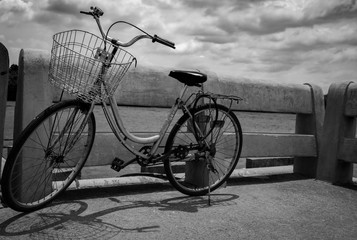 Fototapeta na wymiar Vintage lonely bicycle parked on concrete road by the river with grey sky and clouds. Travel alone. Lonely life. Worthless or abandoned bike. Hopeless, despair, depressed, sad and grief concept.