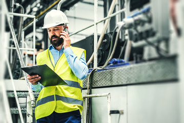 A portrait of an industrial man engineer with smartphone in a factory, working.