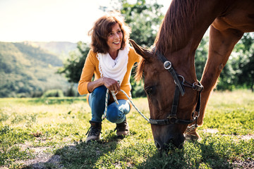 A senior woman crouching and a horse grazing by a stable.