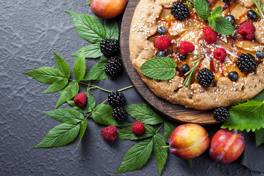 Whole-grain galette with plums and berries on dark background, top view