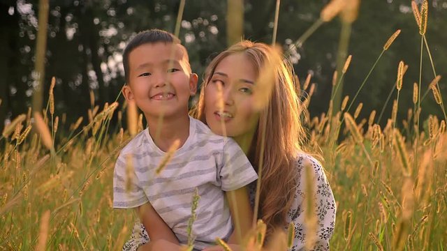 Smiling Asian Mother With Her Little Son Sitting And Hugging In Grass On What Field Landscape