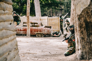 female paintball player in goggle mask and camouflage sitting with paintball gun while her team sitting behind near broken car outdoors