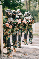 female paintball player holding marker gun with her team in protective masks and camouflage playing paintball outdoors