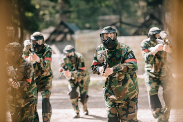 selective focus of paintball team in uniform and protective masks playing paintball with marker guns outdoors