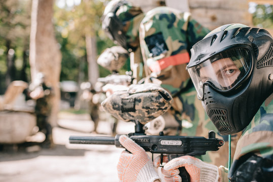 portrait of male paintball player in protective mask holding marker gun and looking at camera outdoors