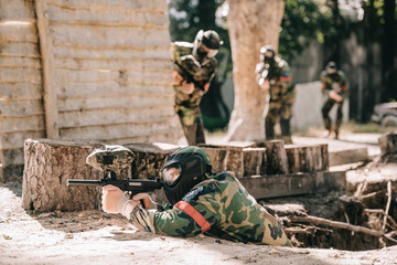 Fototapeta premium selective focus of paintball player laying on ground and aiming with marker gun and his team hiding behind wooden wall outdoors