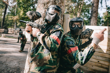 Obraz premium male paintball player in camouflage and goggle mask pointing by finger to his teammate with marker gun standing near tree outdoors