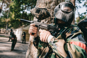 Fototapeta premium paintball players in camouflage and protective masks talking and hiding behind tree outdoors