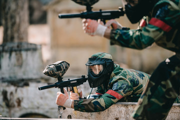 selective focus of paintball player in goggle mask aiming with marker gun near teammate outdoors