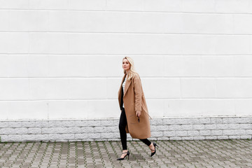 Beautiful young stylish blonde woman wearing long beige coat and black high heel shoes walking...