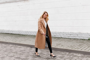 Beautiful young stylish blonde woman wearing long beige coat and black high heel shoes walking...