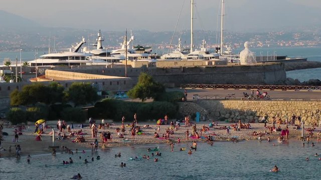 Crowded beach (Plage de la Gravette) near Port Vauban in Antibes, southern France. Boats in harbour, statue "Le Nomade" by Plensa, and hill with residential houses in background. Establishing shot.
