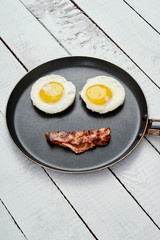 two delicious fried eggs and fried bacon in a pan on a white kitchen table