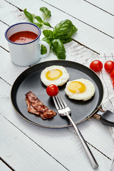 two delicious fried eggs and fried bacon in a pan on a white kitchen table with tomatoes juice and basil leaves