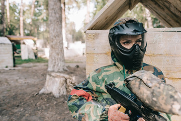 serious female paintballer in protective mask and camouflage uniform standing with paintball gun outdoors