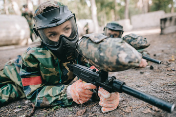 female paintball player in goggle mask and camouflage with marker gun crawling on ground outdoors