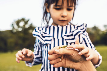 Closeup portrait of cute little girl explores the nature outdoor. Loving mother and daughter spend time together in the park. Pretty Caucasian girl playing with a green grasshopper.