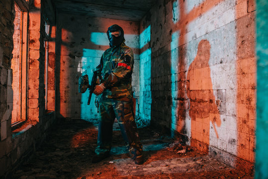 confident male paintball player in goggle mask and camouflage holding paintball gun in abandoned building