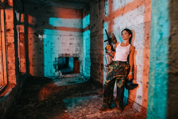 Obraz na płótnie Canvas young female paintballer in camouflage and white singlet holding paintball gun and goggle mask in abandoned building