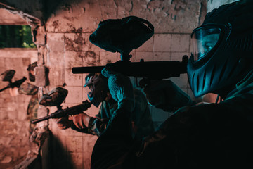 close up view of paintball team in uniform and protective masks playing paintball with marker guns in abandoned building