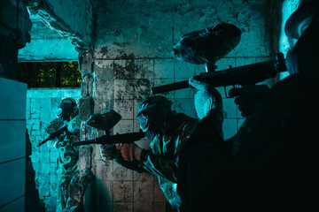 paintball players in uniform and protective masks playing paintball with marker guns in abandoned building