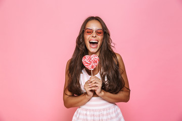 Photo of fashion woman 20s wearing sunglasses laughing and eating lollipop, isolated over pink background - obrazy, fototapety, plakaty