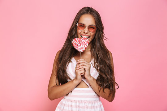 Photo of young woman 20s wearing sunglasses laughing and eating lollipop, isolated over pink background
