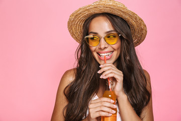 Photo closeup of european woman 20s wearing sunglasses and straw hat drinking juice from glass bottle, isolated over pink background - Powered by Adobe