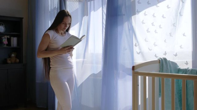 Future young mother between 30 and 35 years old is reading a book close to the open window.