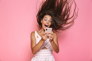 Photo of caucasian woman 20s wearing dress smiling and holding mobile phone with hair shaking, isolated over pink background