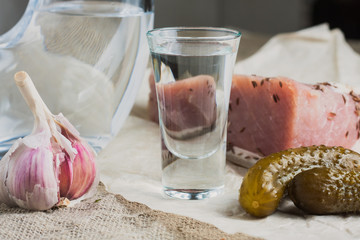 A glass of vodka, bacon, cucumber and garlic on a linen cloth
