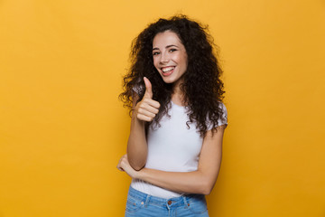 Image of young woman 20s with curly hair smiling and showing thumb up, isolated over yellow...