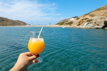 Summer drink cocktail of fresh orange juice with ice in the woman hand on the seascape background of Kolona beach Kythnos island Cyclades Greece.