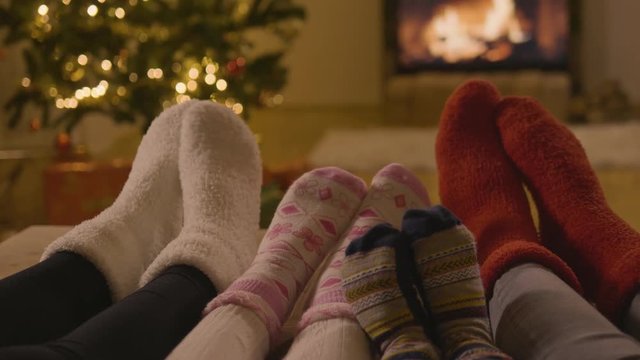Eye level shot of 4 pairs of feet with Christmas socks on in front of the fireplace and the Christmas tree