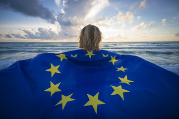 Man holding a fluttering iconic EU flag with circle of stars on beach with stormy turbulent seas in the channel at sunrise