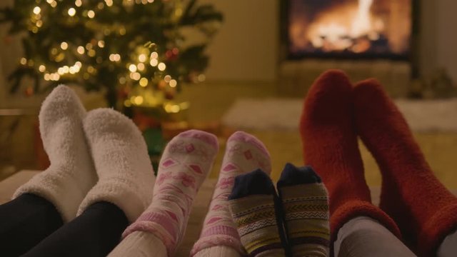 Four pairs of feet with Christmas socks on swinging in front of the fireplace and the Christmas tree