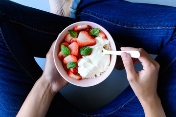 Woman hands holding bowl with cottage cheese, cream, strawberry and mint leaves on gray background. Top view. Healthy lifestyle