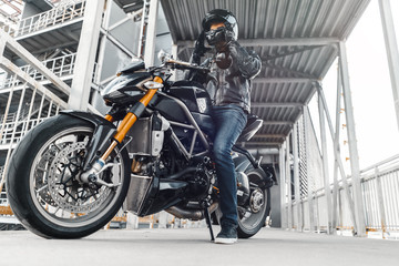 Fototapeta na wymiar Handsome biker sitting on motorcycle and putting on helmet. Urban background, parking.