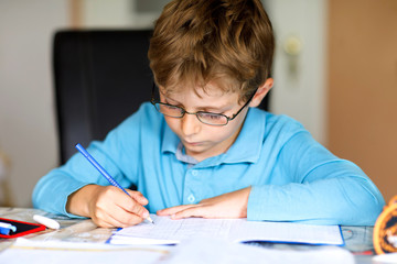 Cute little kid boy with glasses at home making homework, writing letters and doing maths with colorful pens. Little child doing exercise, indoors. Elementary school and education, imagine fantasy