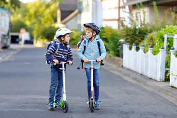 Two school kid boys in safety helmet riding with scooter in the city with backpack on sunny day....