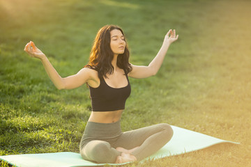 Young girl doing yoga in the park. Woman practicing yoga performing lotus position outdoors with closed eyes. Young attractive slim girl in bodysuit relaxing and doing exercises on a yoga mat. Happy