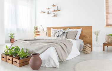 Pillows and sheets on wooden bed in bright bedroom interior with plants and windows. Real photo