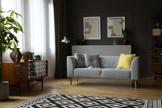 Dark living room interior in real photo with window with curtains, patterned carpet, retro cupboard and grey sofa with cushions