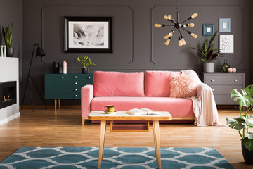 Wooden coffee table with tea cup and open book standing on carpet in dark living room interior with powder pink lounge with fur cushion and blanket