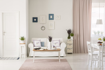 Lavender flowers on table in front of white couch in flat interior with pink drapes and posters....