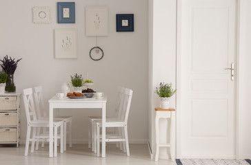 White chairs and table in minimal dining room interior with plants, posters and door. Real photo